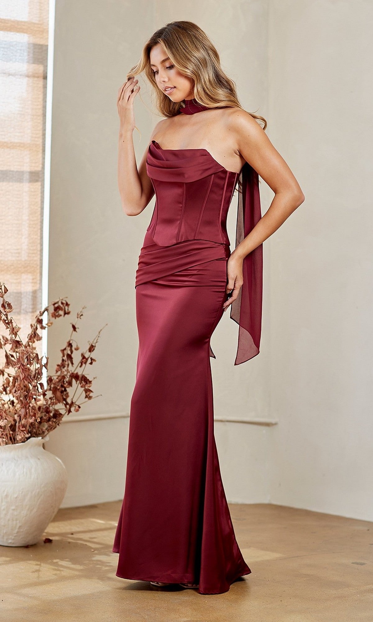 A woman wearing the wine-colored Long Prom Dress: Amelia Couture 6138 with a strapless fitted corset bodice and matching neck scarf stands indoors near a large white vase with dried branches.